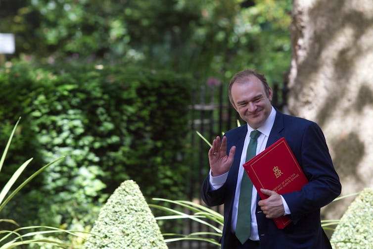 Ed Davey waving while holding his red ministerial folder.