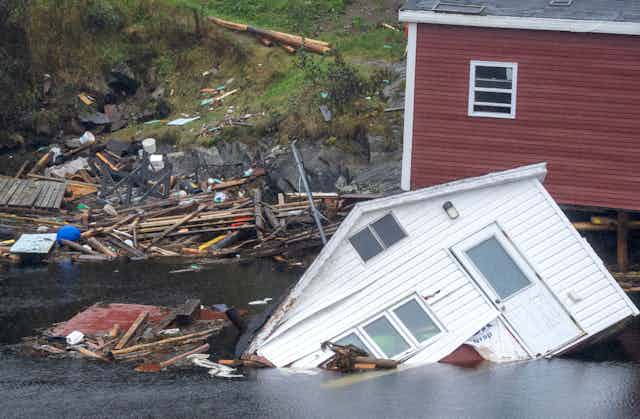 A house sits on its side in the water along a shore.