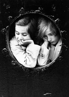 A black and white photo shows two young girls looking through a ship porthole.