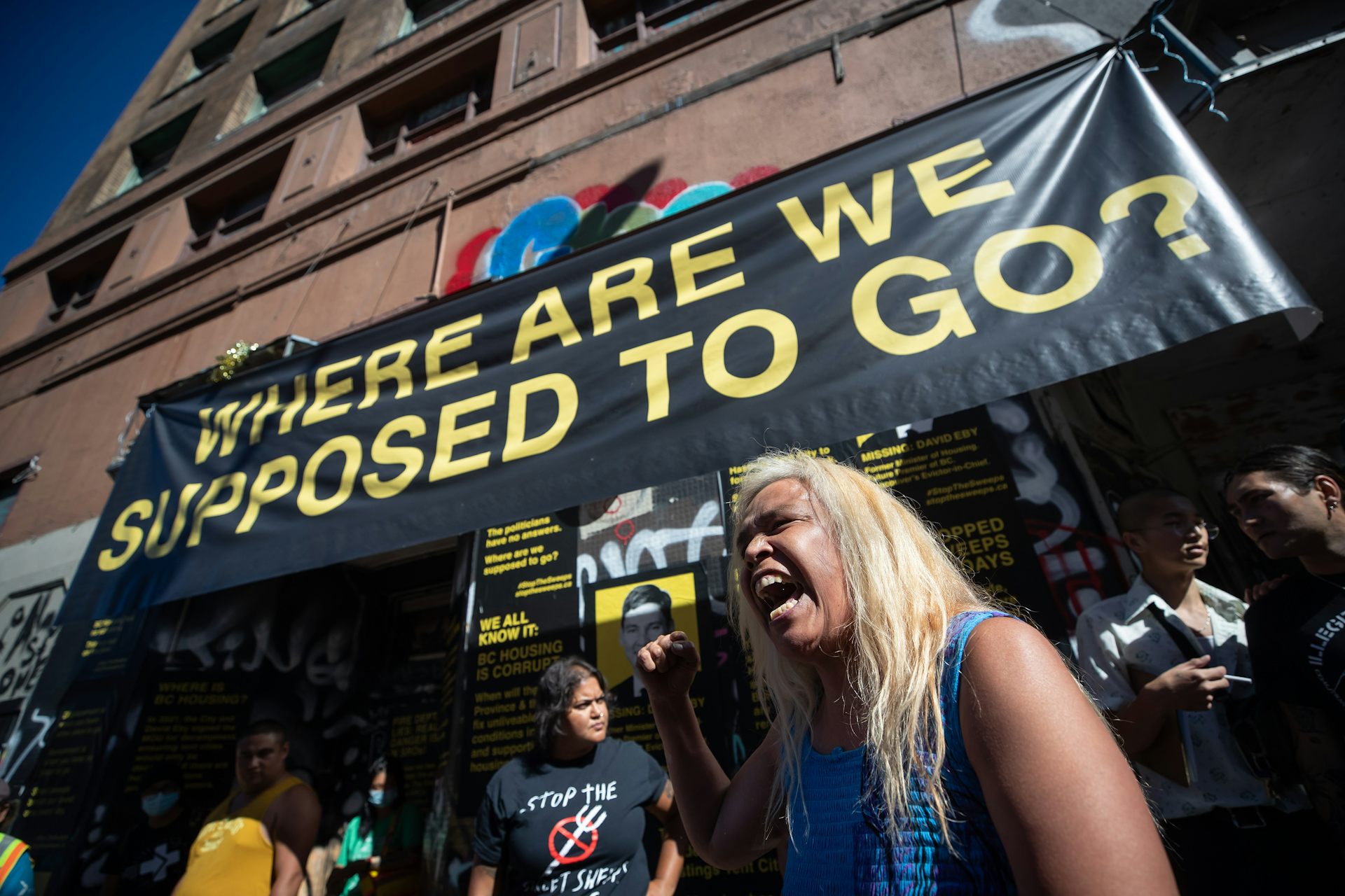A blonde woman shouts to the crowd. A sign above her reads 'where are we supposed to go?'