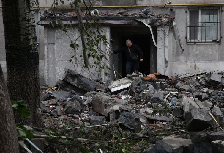A man stands in a doorway of a residential building that has been bombed. Rubble blocks his way.