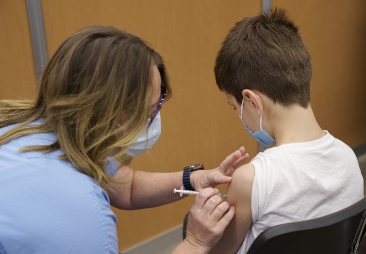 A nurse wearing a mask vaccinates a boy in a white T-shirt also wearing a mask.