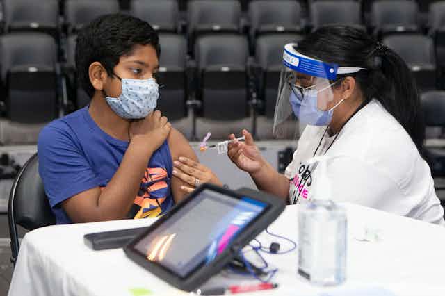 A boy wearing a mask is vaccinated by a woman wearing a face shield.