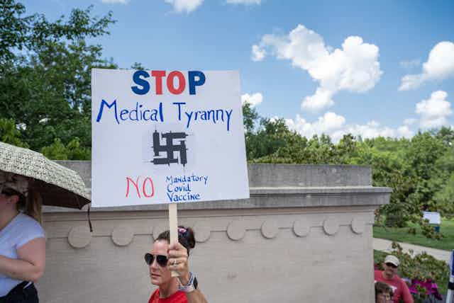 A woman in a red shirt and sunglasses marches with a sign that says, 'Stop medical tyranny, no mandatory covid vaccine' and a swastika.