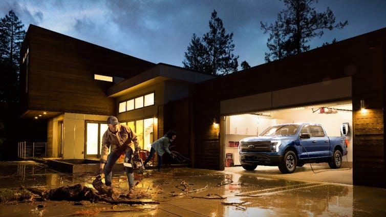 An electric truck parked in a garage, plugged in, while people remove storm debris from a yard