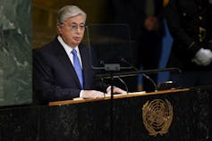 A man with grey hair and glasses in a suit stands before a podium speaking.