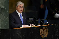 A man with grey hair and glasses in a suit stands before a podium speaking.