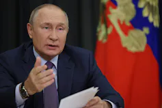 A balding man gestures while speaking, a flag red, blue and gold flag behind him.