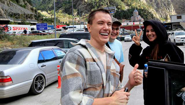 A man smiles and gives a thumb's up while a smiling woman flashes a peace sign. Cars and mountains are seen behind them.