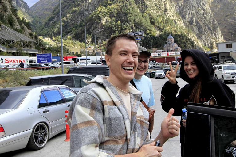 A man smiles and gives a thumb's up while a smiling woman flashes a peace sign. Cars and mountains are seen behind them.