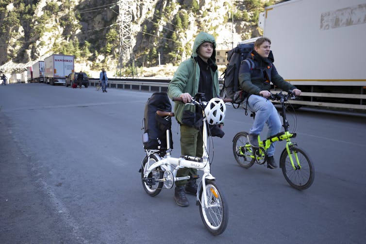 Two smiling young men ride bicycles.