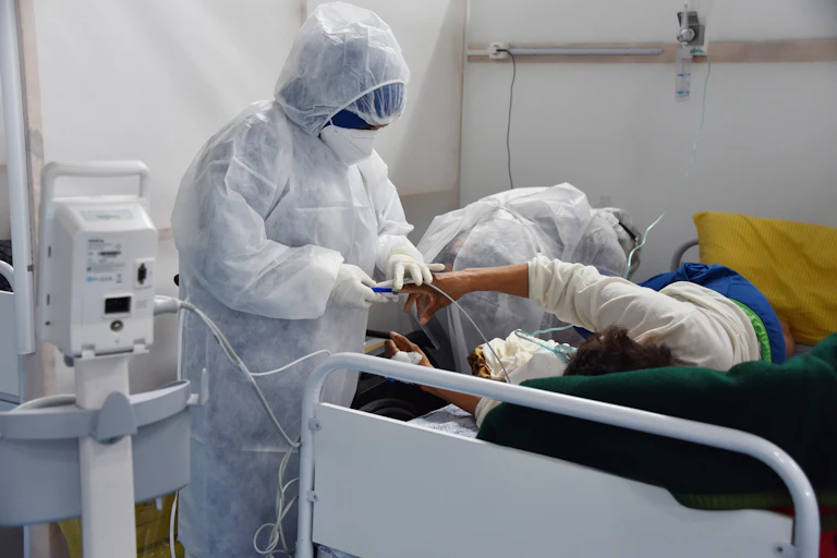A healthcare worker in a full-body white protective suit fits a device to the finger of a person lying on their side in a hospital bed