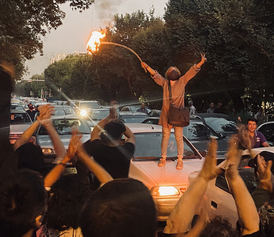A woman stands on the roof of a car with her arms in the air holding a stick with a burning hijab atached.