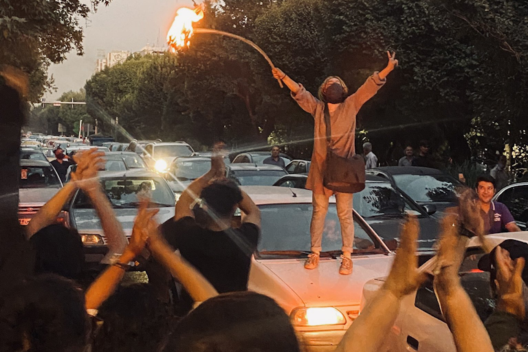 A woman stands on the roof of a car with her arms in the air holding a stick with a burning hijab atached.