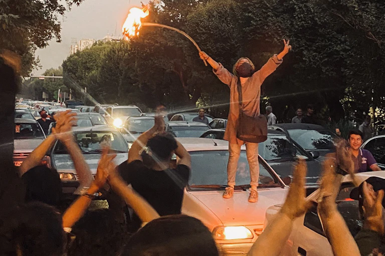 A woman stands on the roof of a car with her arms in the air holding a stick with a burning hijab atached.