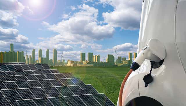 Rows of solar panels and an electric car charging in the foreground with a city skyline and wind turbines in the background.