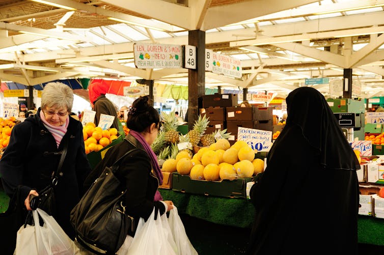 Three women, one in a hijab, with shopping bags at a fruit market.