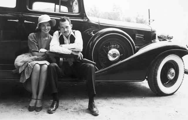 A man with a Gatling gun on his lap and a woman wearing a hat sit on the edge of a classic car