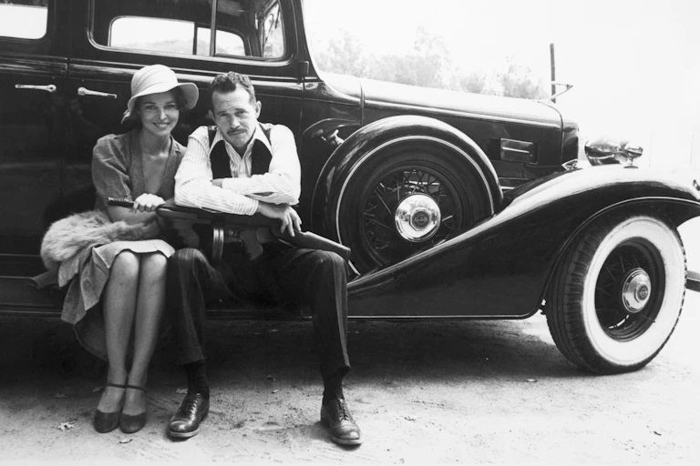 A man with a Gatling gun on his lap and a woman wearing a hat sit on the edge of a classic car