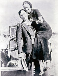 A white man holds a white hat in his right hand and a woman in his left in front of a classic car in a black and white photo