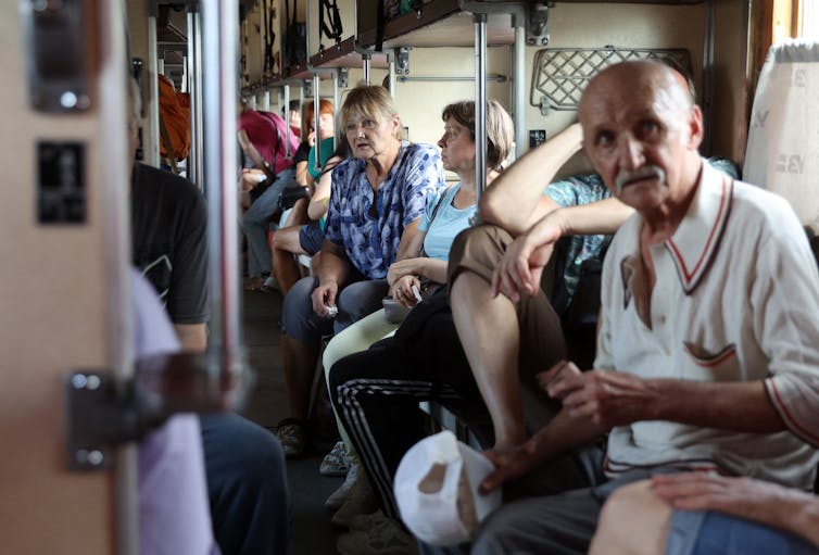 A group of older looking people sit on a train, looking distressed.
