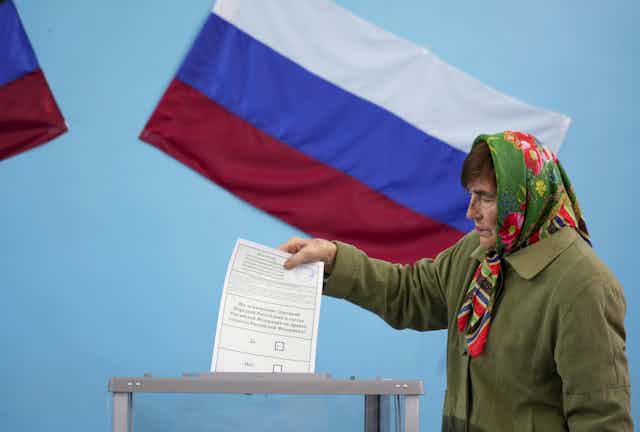 An older woman wearing a green jacket and a colorful babushka around her head appears to drop a white voting ballot into a box, with a Russian flag behind her. 