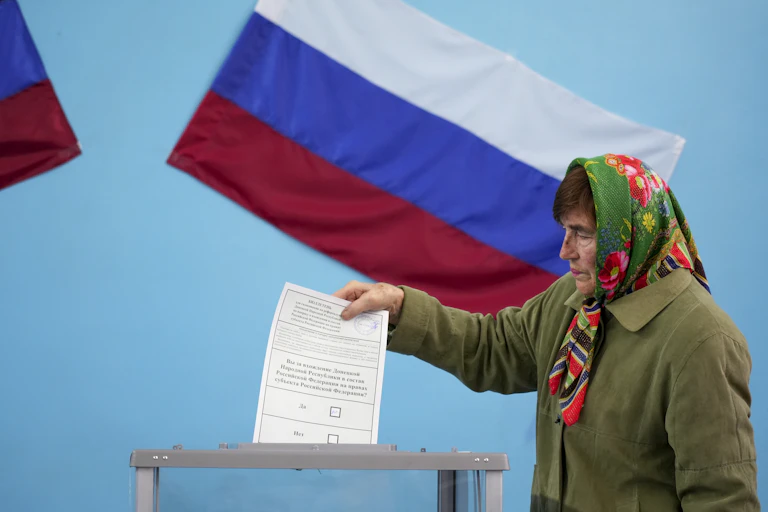 An older woman wearing a green jacket and a colorful babushka around her head appears to drop a white voting ballot into a box, with a Russian flag behind her.