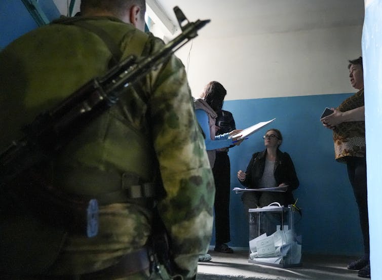 The back of a solider carrying a gun is shown, while a woman in the background appears to vote in a plain room with blue and white walls.