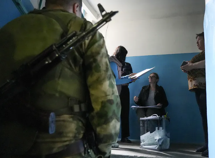 The back of a solider carrying a gun is shown, while a woman in the background appears to vote in a plain room with blue and white walls.