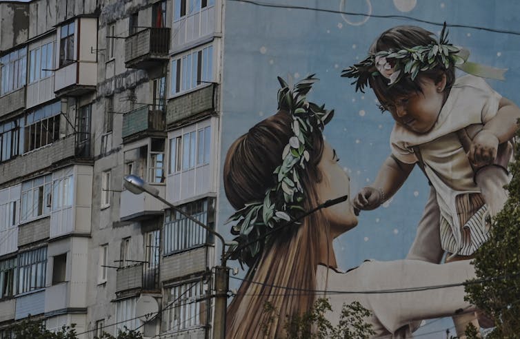 A large mural on the side of a run-down looking building shows the side of a woman holding a baby to the sky, with both baby and woman wearing wreath garlands on their heads.