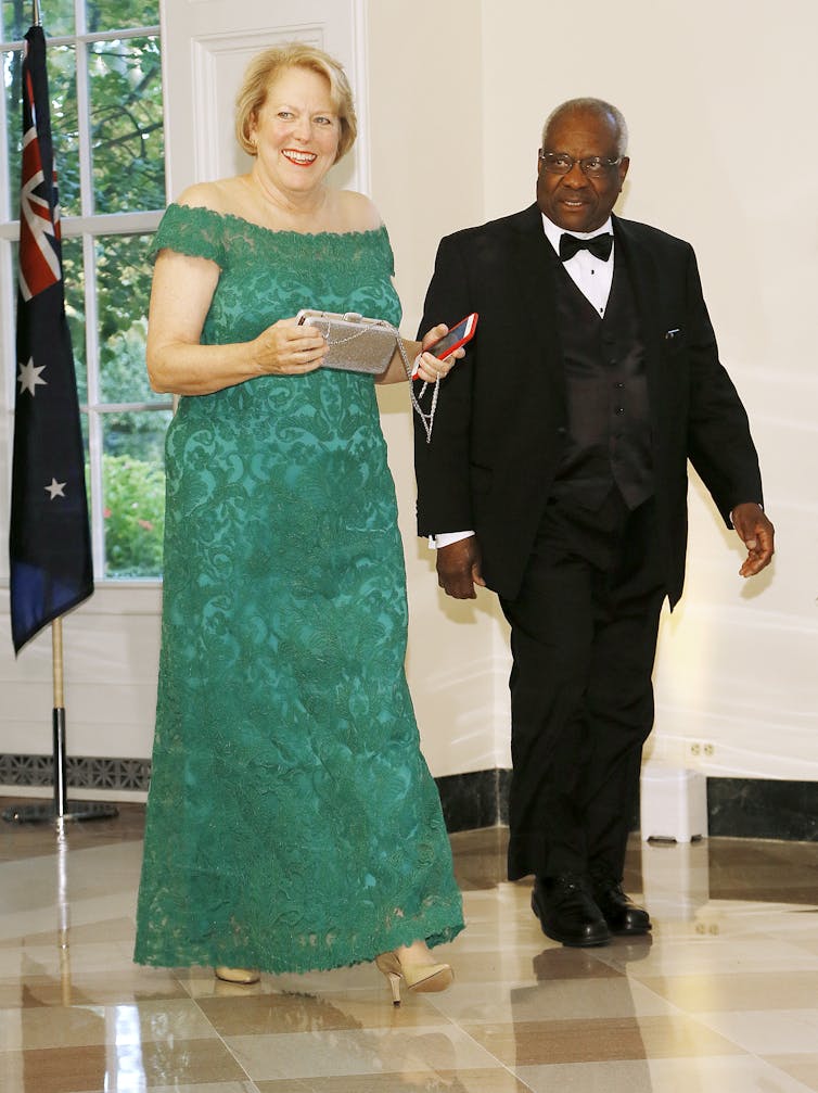 A white womn dressed in a full-length green gown is walking next to a middle-aged Black man wearing a black tuxedo.
