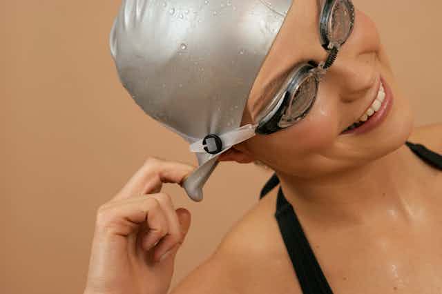 A female swimmer wearing a bathing cap and goggles turns her head sideways to get the water out of her ear.