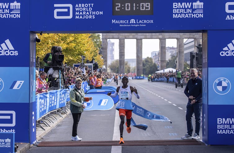 A man running through a blue ribbon with a dark blue gate above him reading 2:01:09