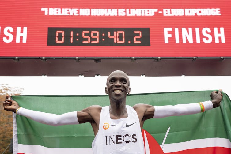 A happy man holding a Kenyan flag stands in front of a timer that reads 1:59:40.2