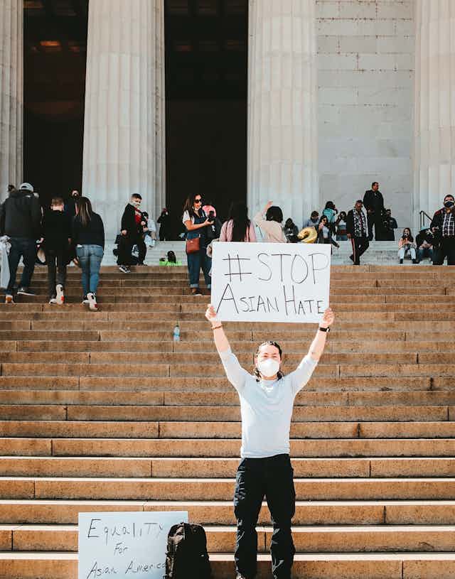 A protestor stands in front of stairs and holds a sign saying '#STOP Asian Hate"