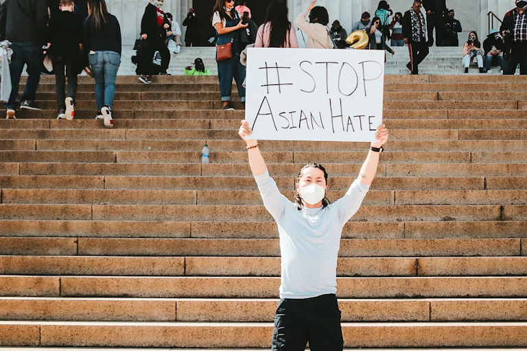 A protestor stands in front of stairs and holds a sign saying '#STOP Asian Hate"