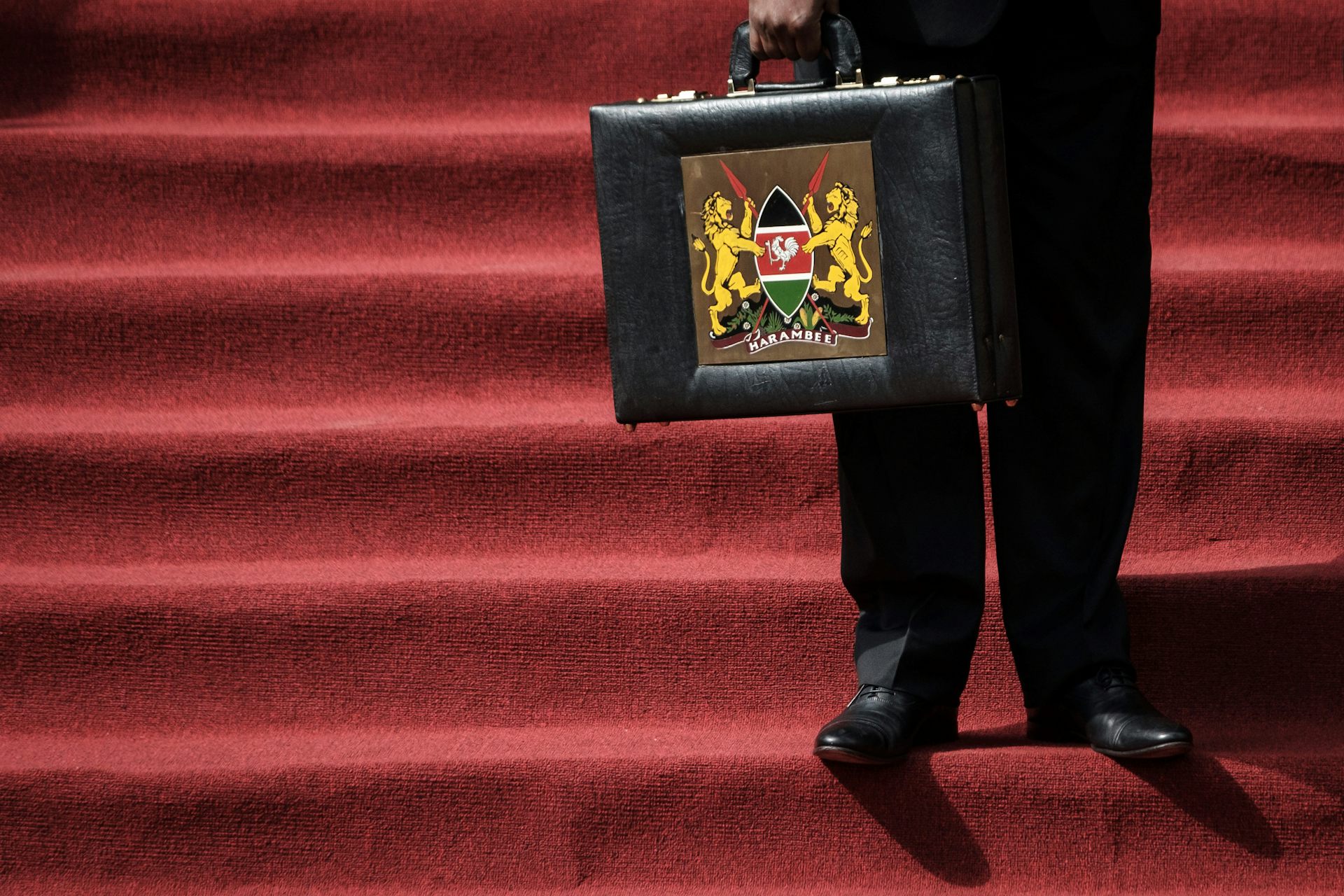 An cropped image showing the feet of a man standing on stairs on a red carpet while holding the Kenyan budget briefcase