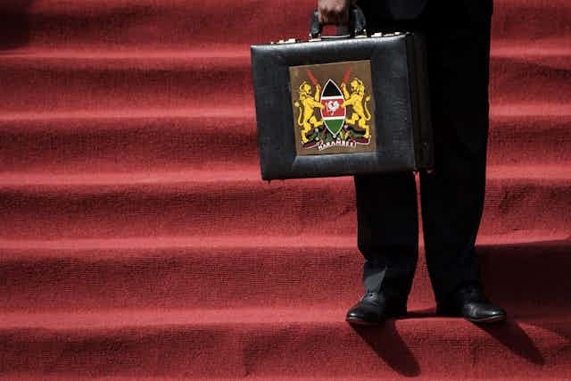 Bottom half of man holding a black briefcase with Kenya's coat of arms on the front.
