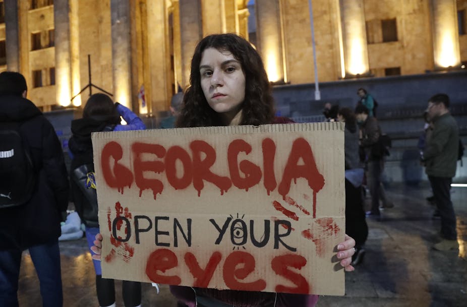 A Georgian woman holds up a signs saying:  Georgia open your eyes