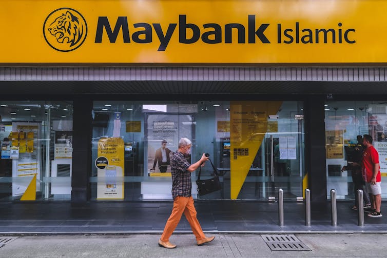a man walks in front of a bank with a yellow sign that reads MAYBANK ISLAMIC