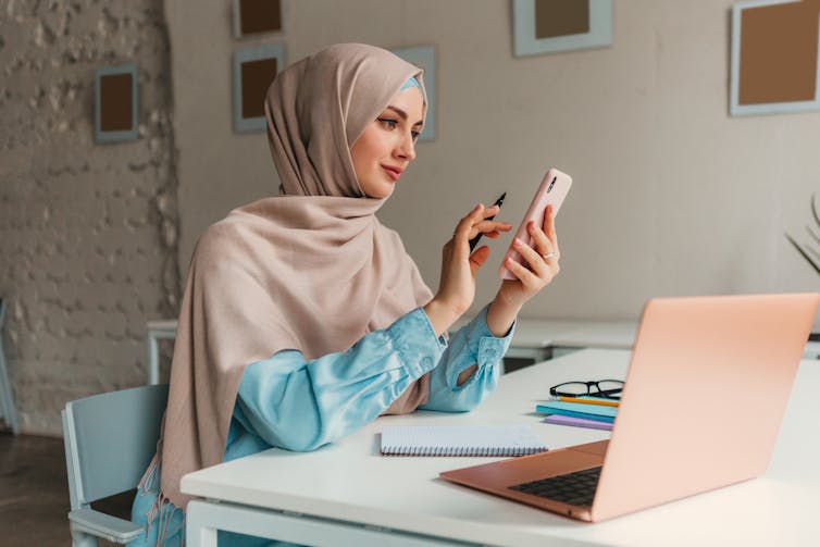 A woman wearing a hijab sits at a desk while using a cell phone. A laptop and notebook are on the desk.