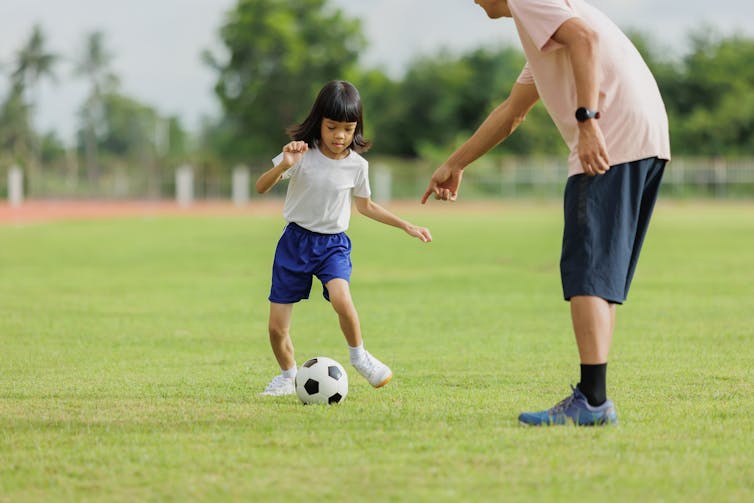 A young girl kicking a soccer ball, with a man partly out of frame pointing at the ball.