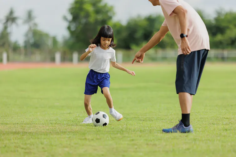 A young girl kicking a soccer ball, with a man partly out of frame pointing at the ball.