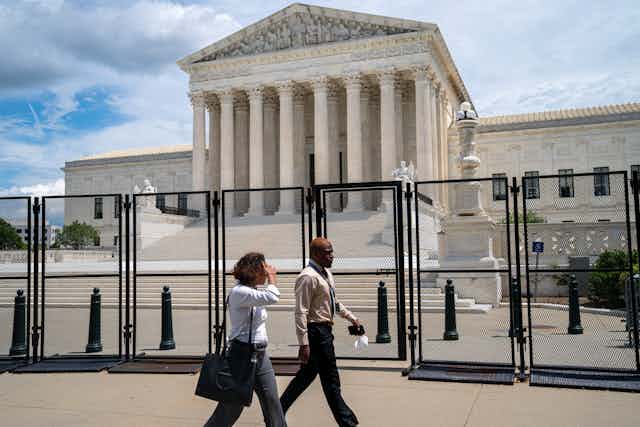 Two people walk past the Supreme Court on a sunny day, with a big fence around it. 
