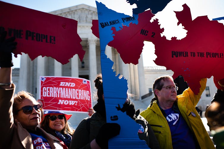 People hold up cut-out blue and red maps of states that say things like 'balance power,' and a sign that says 'end gerrymandering now,' outside the Supreme Court.