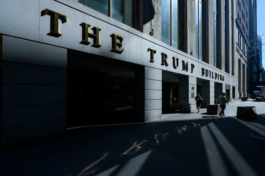 The facade of a building with 'The Trump Building' in embossed letters on it. A couple of people walk by.
