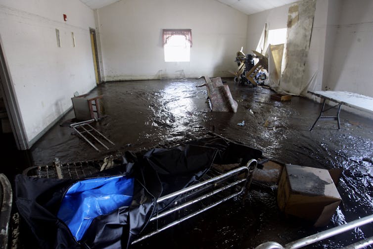 A muddy room with an overturned chair and a gurney. The mud line on the interior wall from floodwater is near the ceiling.