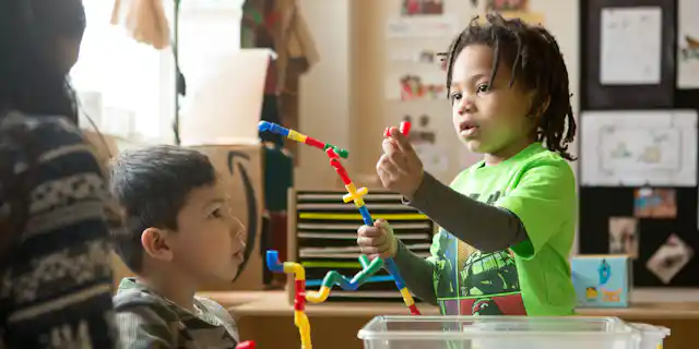 A child holds up loose tubular parts they are building a structure with and a child and educator are looking on.