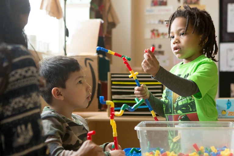 A child holds up loose tubular parts they are building a structure with and a child and educator are looking on.