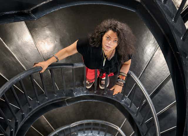 A woman stands on a metal spiral staircase, looking up frankly at the camera. She is dressed in street clothes with a bush of black hair and colourful bracelets.